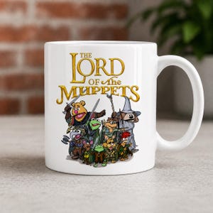 Peut inclure: Mug en céramique blanche avec une illustration colorée de personnages des Muppets dans une parodie du Seigneur des Anneaux. Le mug porte l'inscription "THE LORD OF THE MUPPETS" en lettres dorées.