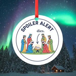 Op de afbeelding: Een ronde kerstversiering met de tekst "SPOILER ALERT dies." en een illustratie van de kerststal. De ornament heeft een rood lint om op te hangen, tegen een besneeuwde bosachtergrond.