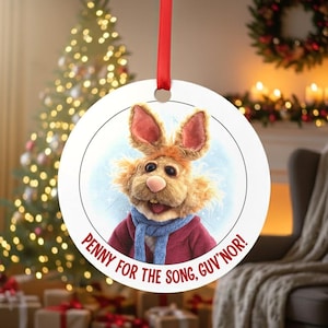 Könnte beinhalten: Runder Weihnachtsschmuck mit einer Cartoon-Kaninchenfigur mit rotem Mantel und blauem Schal. Der Schmuck trägt den Text "Penny for the song, guv'nor!" in Rot. Ein rotes Band ist zum Aufhängen befestigt.