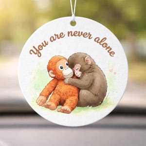May include: Circular white ornament with the words "You are never alone" in brown script. It shows a watercolour illustration of two hugging monkeys, one orange and one brown, set against a grassy backdrop.