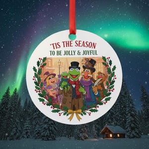 May include: A circular Christmas decoration with a red ribbon. The ornament displays a festive illustration of the Muppets in Victorian clothing, framed by a green wreath and the text "'Tis the Season to be Jolly & Joyful."