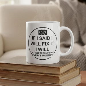 Könnte beinhalten: Weiße Keramiktasse mit dem Text "IF I SAID I WILL FIX IT I WILL NO NEED TO REMIND ME EVERY 6 MONTHS!" in Schwarz. Die Tasse steht auf einem Stapel Bücher.