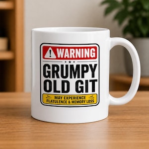 May include: White ceramic mug with a black and red warning label that reads "WARNING GRUMPY OLD GIT" with the text "MAY EXPERIENCE FLATULENCE & MEMORY LOSS". The mug has a white handle and is on a wooden surface.