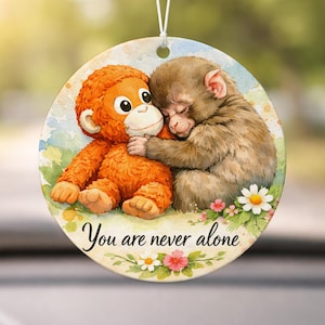 May include: Circular ornament with a watercolour illustration of two monkeys. One is orange and hugging the other, which is brown and sleeping. The text "You are never alone" is written below the monkeys, surrounded by flowers.