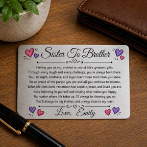 Könnte beinhalten: Eine rechteckige silberfarbene Metall-Geldbörsenkarte mit dem schwarzen Aufdruck 'Sister To Brother', einer sentimentalen Botschaft und der Signatur 'Love, Emily'. Die Karte ist mit kleinen rosa und violetten Herzillustrationen verziert.