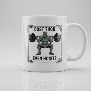 May include: White ceramic mug featuring a cartoon knight lifting a barbell. The knight is in green armor. The mug has the text "DOST THOU EVEN HOIST?" in black, with decorative borders.