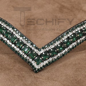 May include: Close-up of a decorative horse bridle browband. The browband features a V-shaped design with alternating rows of emerald green and clear crystal rhinestones. The rhinestones are set on a black background.