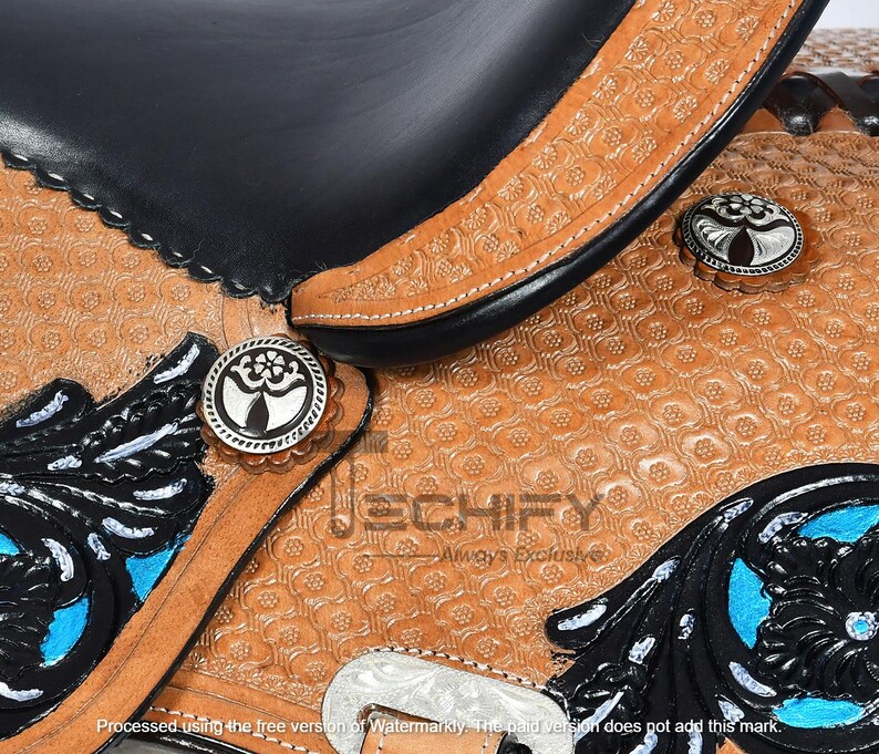 May include: Close-up of a western saddle with intricate tooling and decorative silver conchos. The saddle features black leather accents, turquoise details, and a floral pattern embossed on the tan leather. The saddle is a detailed piece of equestrian equipment.