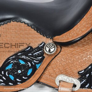 May include: Close-up of a Western saddle with a black seat and brown leather detailing. The saddle features intricate floral designs in black and turquoise, with silver-toned conchos and hardware. The leather has a textured pattern.