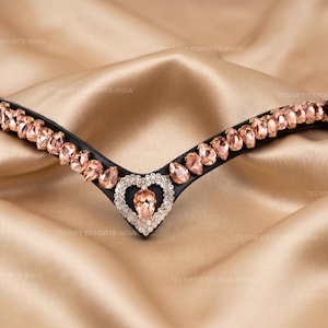 May include: Black leather horse bridle browband adorned with peach-colored gemstones and a heart-shaped centerpiece. The browband is set against a backdrop of a golden satin fabric, highlighting the sparkle of the crystals.