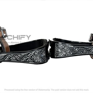 May include: Pair of black and silver Western stirrups with intricate floral designs. The stirrups have a black interior and are accented with silver detailing. Brown leather straps and metal hardware complete the design. The word "CHIFY" is visible.