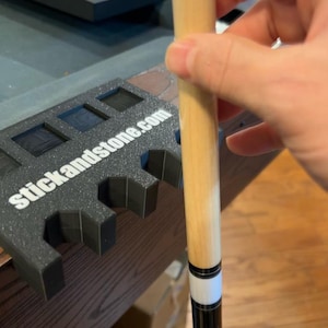 May include: A pool cue with a light wood shaft and a black and white accent. The cue is held near a black pool cue holder with the text "stickandstone.com". The background includes a pool table and a control panel.