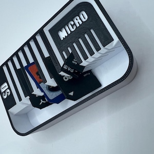 May include: A black and white 3D-printed SD and Micro SD card holder. The holder has slots for multiple cards and is labeled with the words "SD" and "MICRO". Several SD and Micro SD cards are visible inside the holder.