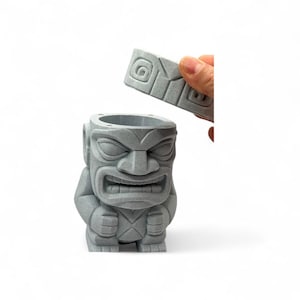 May include: A gray tiki statue with a removable lid. The statue has a detailed carved face with an open mouth and is in a standing position. The lid is being held up by a hand. The statue is made of a textured material.