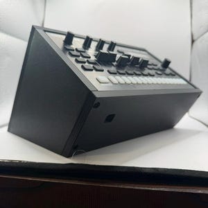May include: A black electronic music production device with numerous knobs, buttons, and sliders. The device has a rectangular shape with a flat top surface and a series of white and black controls. The device is designed for creating and manipulating sounds.