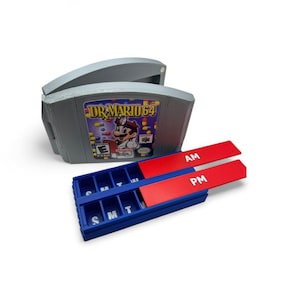 May include: A gray plastic pill organizer shaped like a Dr. Mario 64 video game cartridge. The organizer has a blue base with AM/PM red lids and labeled compartments for each day of the week.