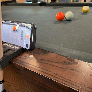 May include: A smartphone records a game of pool, held in a black bracket labeled "MR BIG STUFF". The phone's screen shows the pool balls. Three pool balls are visible on the green felt table: red, white, and yellow.