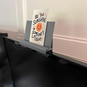 May include: A gray monitor stand holding a card with the text "BE THE SUNSHINE IN SOMEONE'S LIFE TODAY" and a smiling sun graphic. The stand is placed on top of a black computer monitor.