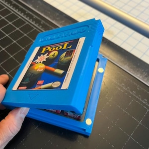 May include: A blue plastic case holding a video game cartridge. The cartridge label reads "Pool" with a graphic of pool balls and cues. The case is open, revealing the game cartridge inside. The background is a cutting mat.