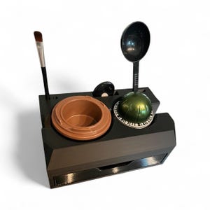 Puede incluir: Un organizador negro con un cajón, que contiene un recipiente marrón, una esfera verde, una cuchara negra, un pincel de maquillaje y un pequeño disco negro. El organizador está hecho de un material oscuro y tiene un diseño moderno.