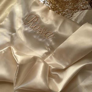 Pode incluir: Uma fronha de cetim em um tom creme suave, bordada com o nome "Rosa" em um delicado script. A fronha est&aacute; dobrada, mostrando o tecido liso e brilhante. O fundo inclui um espelho decorativo e flores secas.