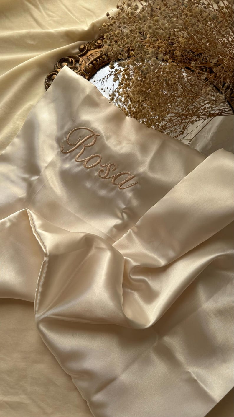 Pode incluir: Uma fronha de cetim em um tom creme suave, bordada com o nome "Rosa" em um delicado script. A fronha est&aacute; dobrada, mostrando o tecido liso e brilhante. O fundo inclui um espelho decorativo e flores secas.