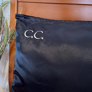 Pode incluir: Uma fronha de cetim azul escuro com as iniciais "C.C." bordadas em branco. A fronha tem uma textura lisa e brilhante. Ao fundo, uma cabeceira de madeira.
