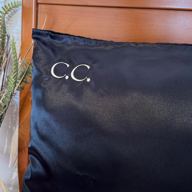Pode incluir: Uma fronha de cetim azul escuro com as iniciais "C.C." bordadas em branco. A fronha tem uma textura lisa e brilhante. Ao fundo, uma cabeceira de madeira.