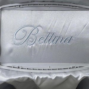 Pode incluir: Tecido de cetim azul claro bordado com o nome "Bettina" em uma fonte cursiva delicada. O bordado &eacute; feito com linha azul clara combinando. O tecido &eacute; esticado dentro de um bastidor de bordado retangular.