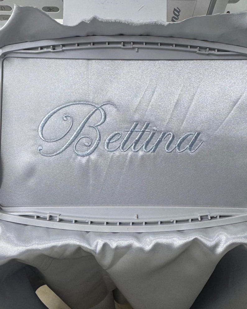 Pode incluir: Tecido de cetim azul claro bordado com o nome "Bettina" em uma fonte cursiva delicada. O bordado &eacute; feito com linha azul clara combinando. O tecido &eacute; esticado dentro de um bastidor de bordado retangular.