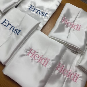 Pode incluir: V&aacute;rios roup&otilde;es de cetim branco dobrados, alguns bordados com o nome "Ernst" em fio azul marinho, e outros com "Heidi" em rosa. Os roup&otilde;es est&atilde;o dispostos sobre uma superf&iacute;cie castanha clara, sugerindo um tema de presente personalizado ou de casamento.