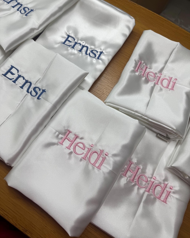 Pode incluir: V&aacute;rios roup&otilde;es de cetim branco dobrados, alguns bordados com o nome "Ernst" em fio azul marinho, e outros com "Heidi" em rosa. Os roup&otilde;es est&atilde;o dispostos sobre uma superf&iacute;cie castanha clara, sugerindo um tema de presente personalizado ou de casamento.