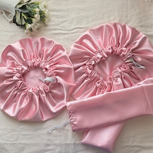 Op de afbeelding: Twee roze satijnen haarbonnets met elastiekjes en een bijpassend roze satijnen slaapmasker.