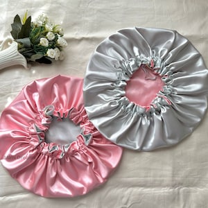 Pode incluir: Dois bonés de cabelo de cetim, um rosa e outro prateado, com forro branco. Os bonés são franziados na parte superior e têm um cordão.