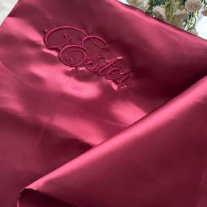 Pode incluir: Tecido de cetim bord&ocirc; com a palavra "Ester" bordada em fonte cursiva. O tecido est&aacute; dobrado, criando sombras e reflexos. O tema geral &eacute; luxo e personaliza&ccedil;&atilde;o.