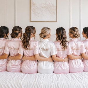 Puede incluir: Siete mujeres con batas de satén, seis en rosa claro y una en blanco. Las batas rosas dicen "Bridesmaid" en letras doradas en la espalda, mientras que la bata blanca dice "Bride". Están de pie sobre una cama blanca.