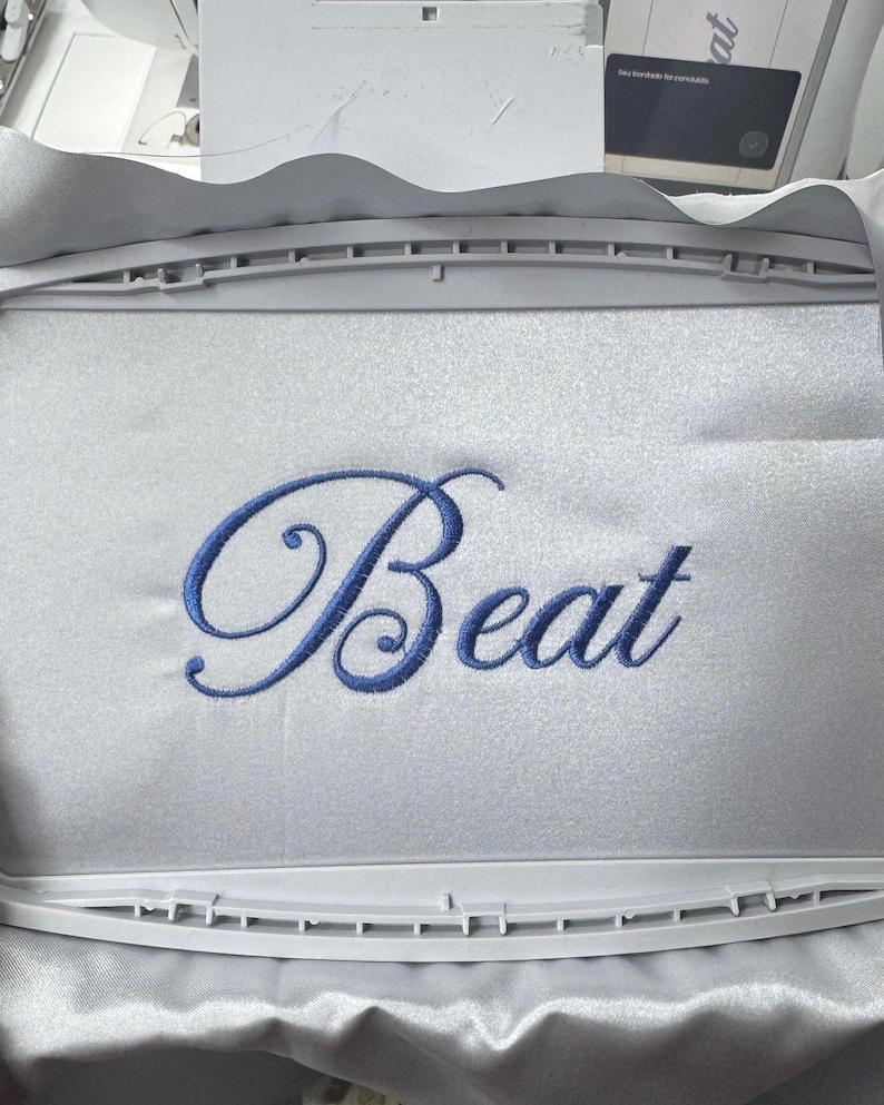 Pode incluir: Tecido branco com a palavra "Beat" bordada em azul. O tecido &eacute; mantido em um bastidor de bordado retangular. A m&aacute;quina de bordar &eacute; vis&iacute;vel ao fundo.
