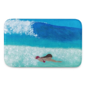 May include: A rectangular bath mat featuring an ocean scene. A swimmer in a red bikini is in the water, with a large blue wave overhead. The water is a mix of turquoise and white, creating a vibrant coastal theme.