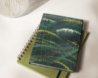 May include: Two notebooks, one spiral-bound and one with an elastic closure, are stacked on a white surface. The top notebook has a green, yellow, and white abstract design. The bottom notebook is a solid olive green color.