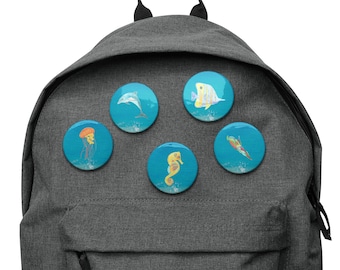 May include: A gray backpack with five round ocean-themed buttons attached. The buttons feature illustrations of a dolphin, seahorse, fish, jellyfish, and a colorful sea creature, all set against a blue background.