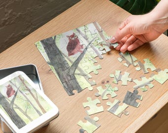 May include: A partially assembled jigsaw puzzle featuring an owl perched on a tree branch in a forest scene. The puzzle pieces are scattered on a wooden surface, with a metal tin containing the puzzle image visible.