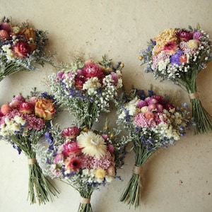Puede incluir: Seis pequeños ramos de flores secas que presentan una mezcla de flores rosas, moradas, amarillas y blancas. Cada ramo está atado con cuerda en la base y contiene una variedad de tallos, pétalos y pequeñas flores de relleno secas.
