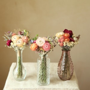 May include: Three glass vases containing dried flower bouquets. The left vase is clear and smooth, the center vase is clear with a diamond-textured pattern, and the right vase is tinted mauve with a textured surface, all holding colorful dried blooms.
