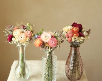 May include: Three glass vases containing dried flower bouquets. The left vase is clear and smooth, the center vase is clear with a diamond-textured pattern, and the right vase is tinted mauve with a textured surface, all holding colorful dried blooms.