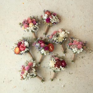 May include: Seven small bouquets of dried flowers in shades of pink, white, and orange are arranged on a textured, light-colored surface. Each bouquet is wrapped with twine. The flowers appear to be a mix of different varieties.