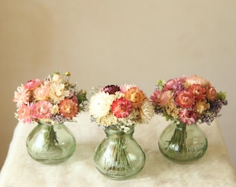 May include: Three small, round, light green glass vases, each containing a dense bouquet of dried flowers in shades of pink, orange, cream, and purple. The glass vases have a short neck and a wide base, displaying a clear, smooth texture.