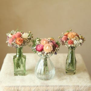 Puede incluir: Tres floreros de vidrio con arreglos de flores secas en tonos rosa, naranja, crema y blanco. Los floreros varían en forma, con dos botellas de vidrio verde altas y delgadas y un florero de vidrio transparente acanalado más ancho.