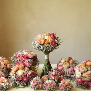 Puede incluir: Una colección de ramos de flores secas con una mezcla de flores rosas, naranjas, amarillas y blancas acentuadas con pequeñas flores de relleno. Los tallos están atados con cordel, mostrando una variedad de cabezas florales texturizadas.
