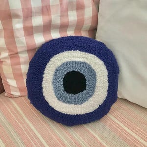 May include: A blue and white evil eye pillow with a black pupil. The pillow is round and has a fluffy texture. It is sitting on a pink and white striped surface.