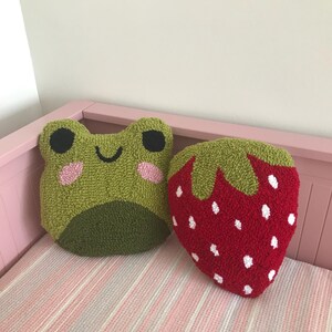 Puede incluir: Dos almohadas de felpa, una con forma de rana verde con una cara sonriente y la otra con forma de fresa roja con semillas blancas.